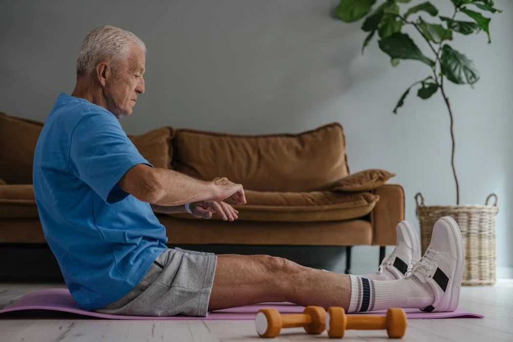 Elderly man in blue shirt exercising on a yoga mat indoors with dumbbells in view.